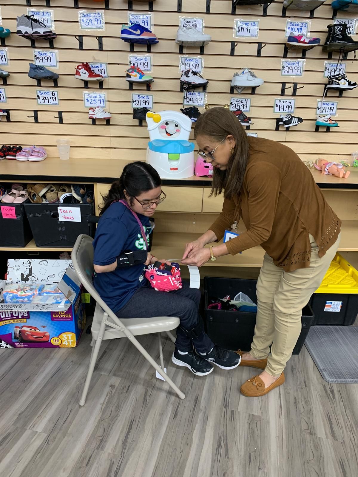 Woman helping person seated on a chair examine an item in a store. Shoes are on display in the background.