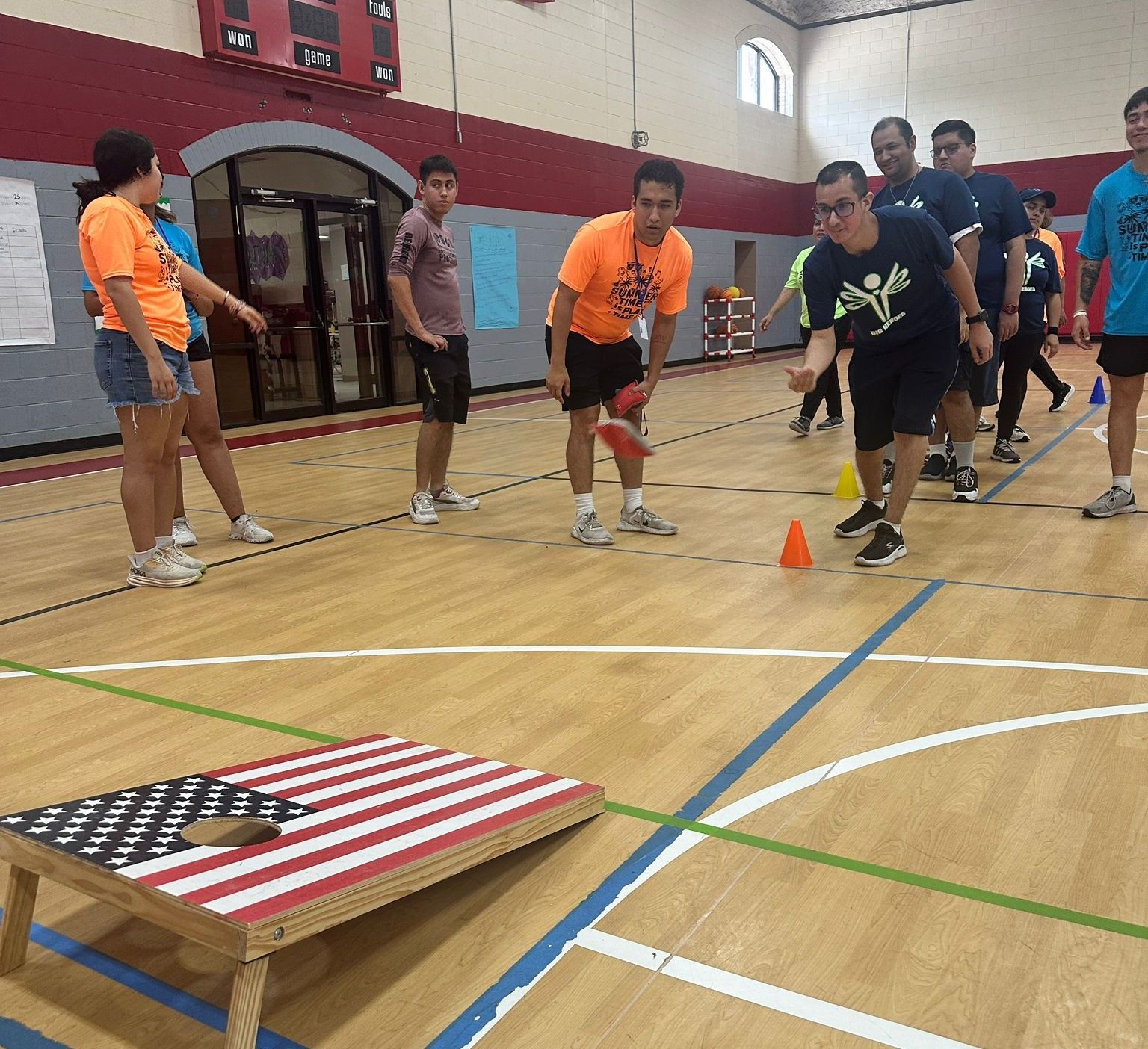 People playing bean bag toss in a gym, one person throwing towards an American flag-themed board.