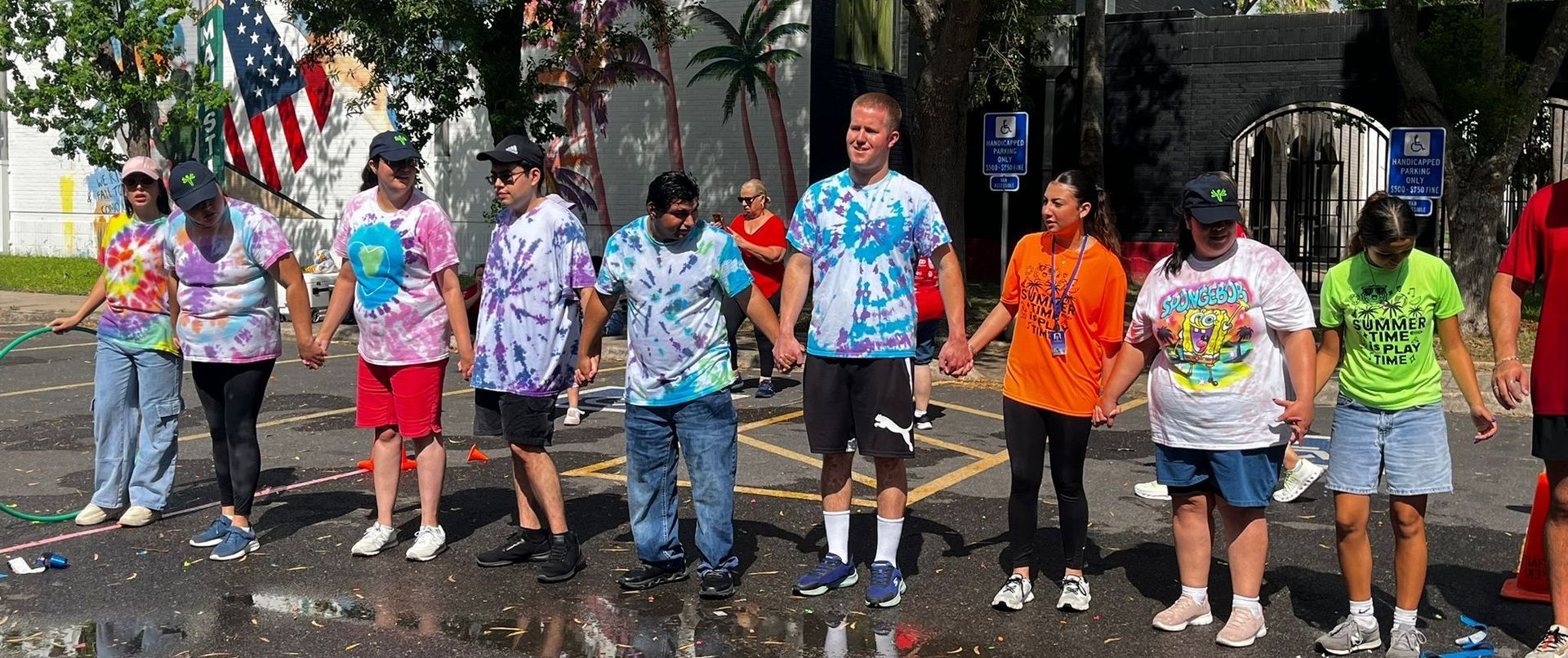 A group of people in tie-dye shirts hold hands in a line outdoors. An American flag is in the background.