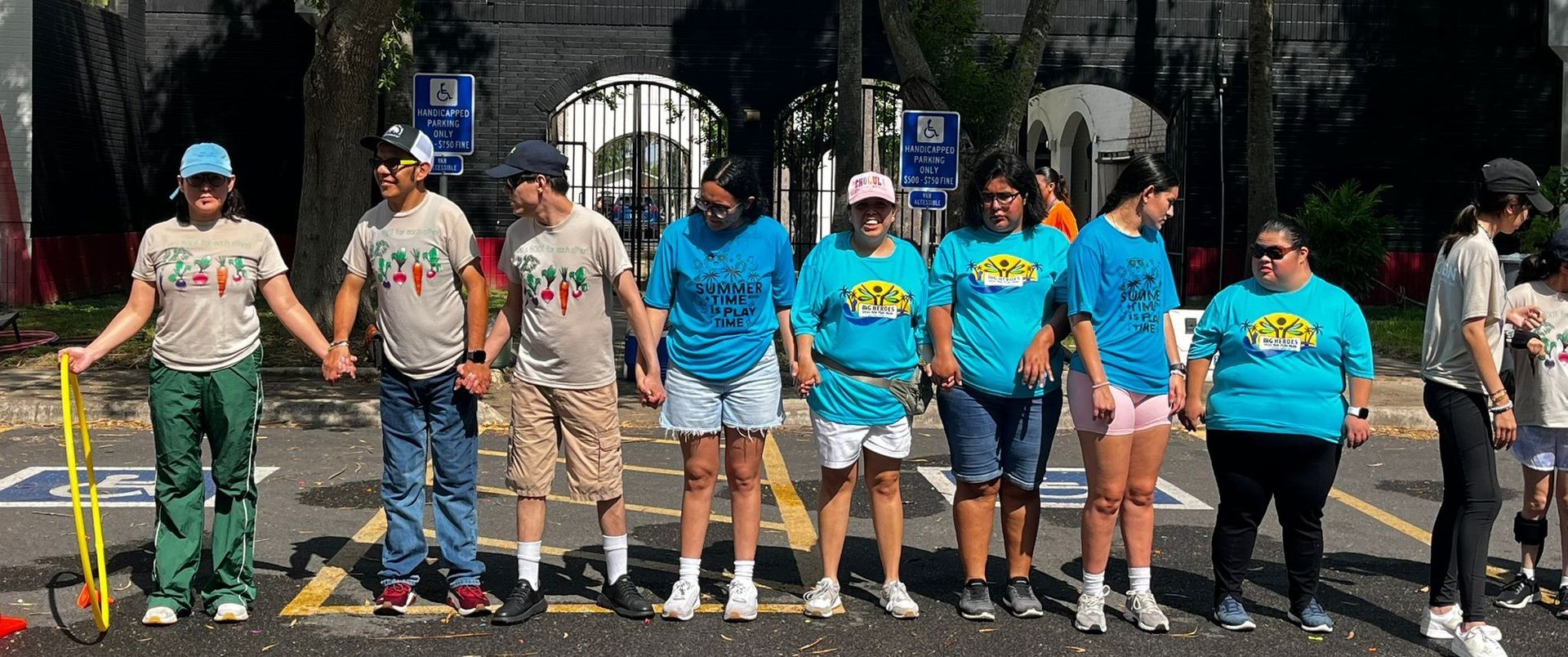 A group of people holding hands in a line, some in blue t-shirts, outdoors in front of a building.