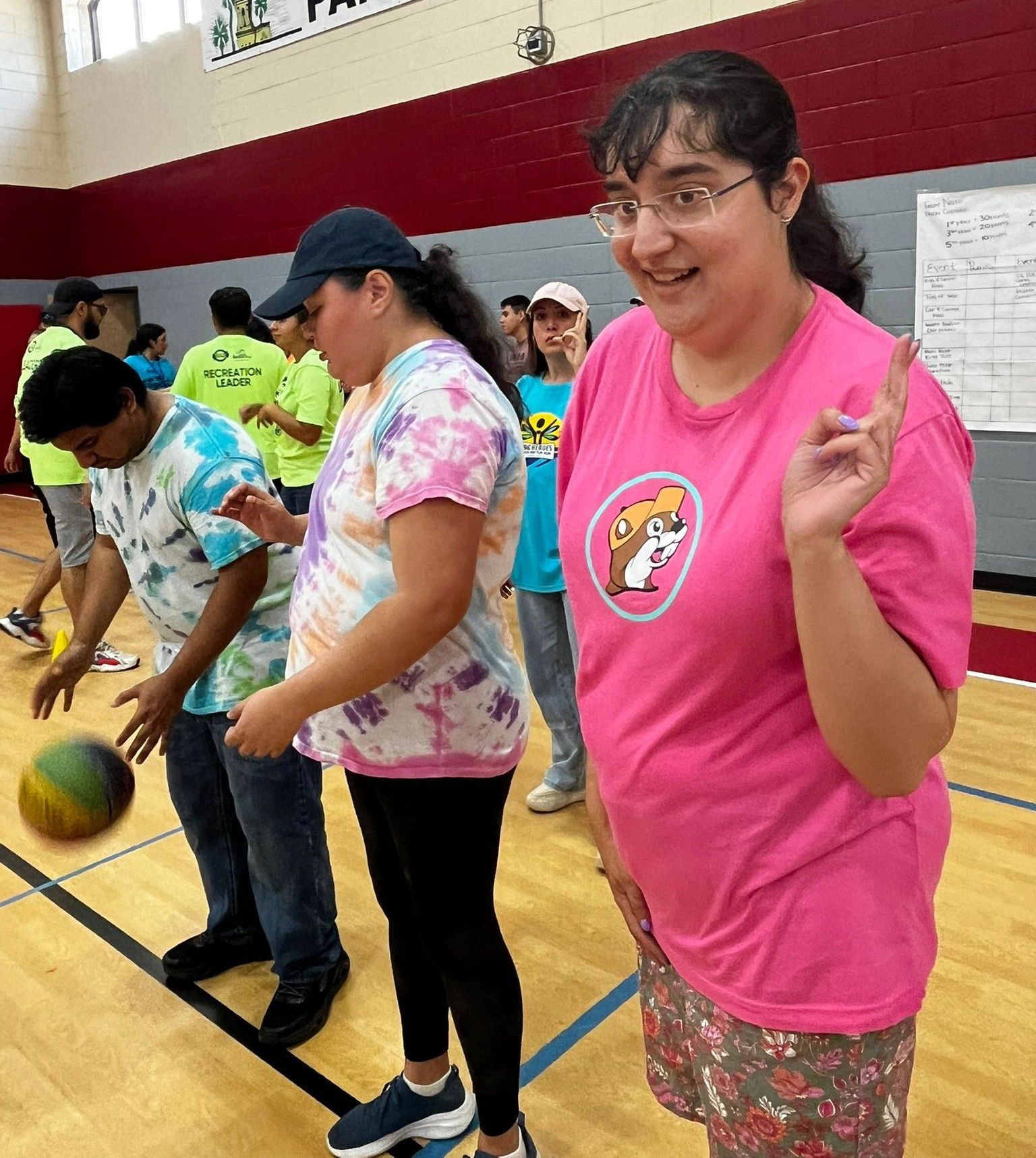 People playing with a ball in a gym, smiling and wearing casual clothes. Woman in pink shirt making a peace sign.