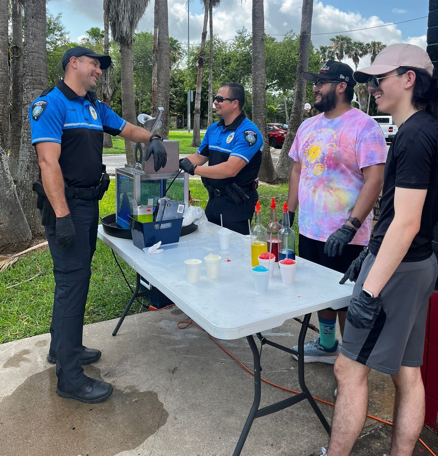 Two police officers serve shaved ice to two young men at an outdoor event.