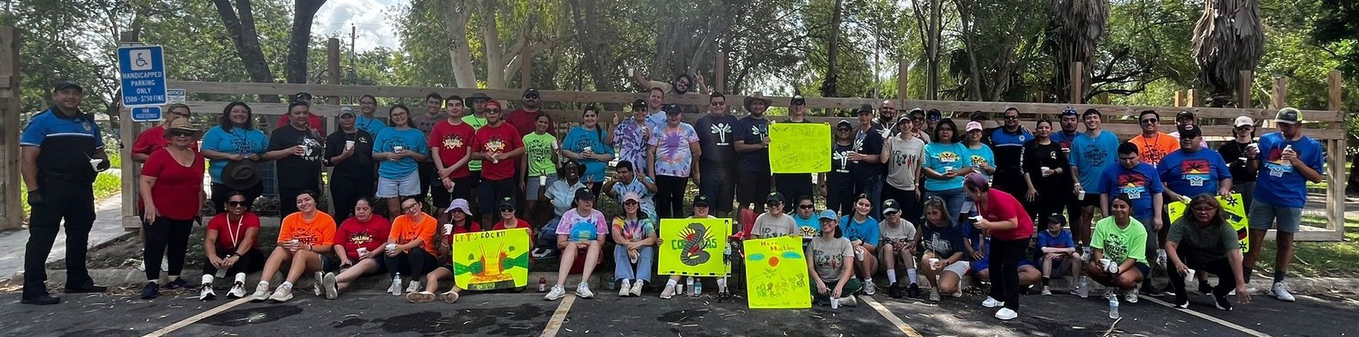 Group of people smiling in an outdoor setting. Some are holding signs, while others are in different colored shirts.