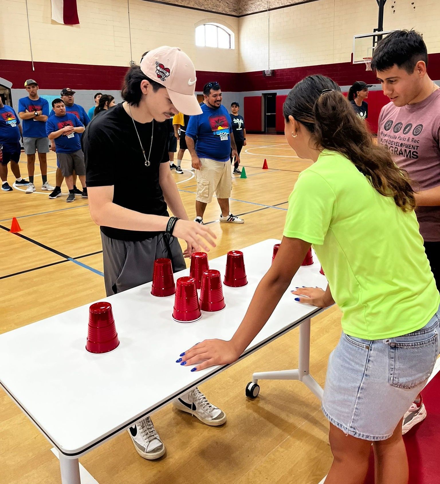 Young people playing a cup-stacking game indoors, two actively participating, others watching.