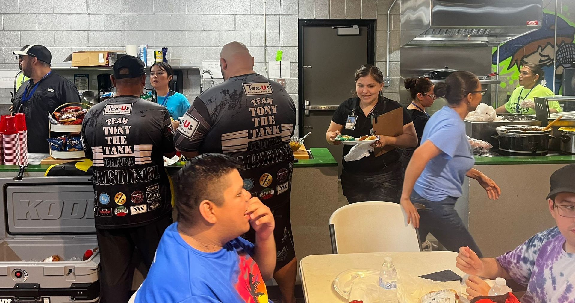 People in a kitchen, some wearing aprons, preparing food. Others sit at tables, possibly at a community event.