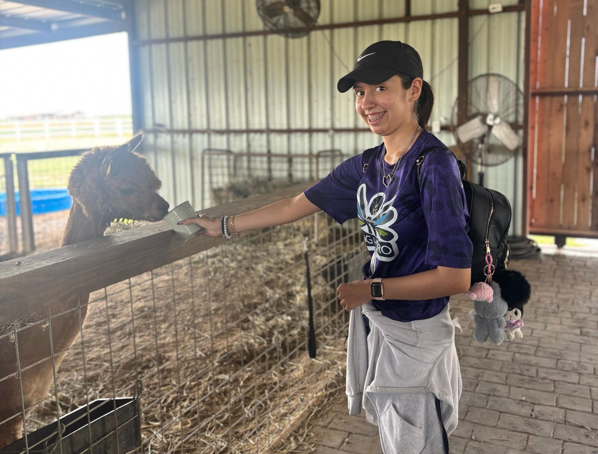 Woman petting an alpaca at a farm. She's smiling and wearing a cap. Barn with hay in the background.