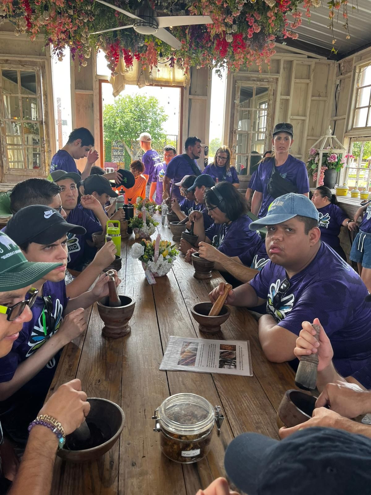 Group in purple shirts pounding in mortars at a table; flowers overhead, open doorway.