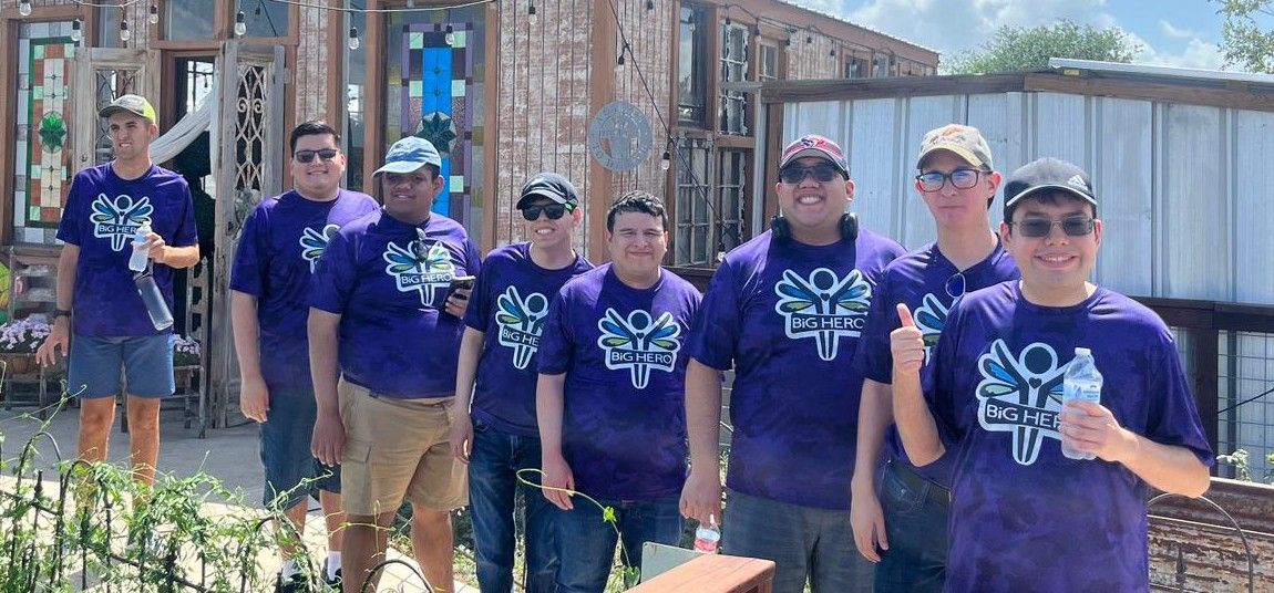 Group of men in purple shirts with logo, posing outside a building, some giving thumbs up.