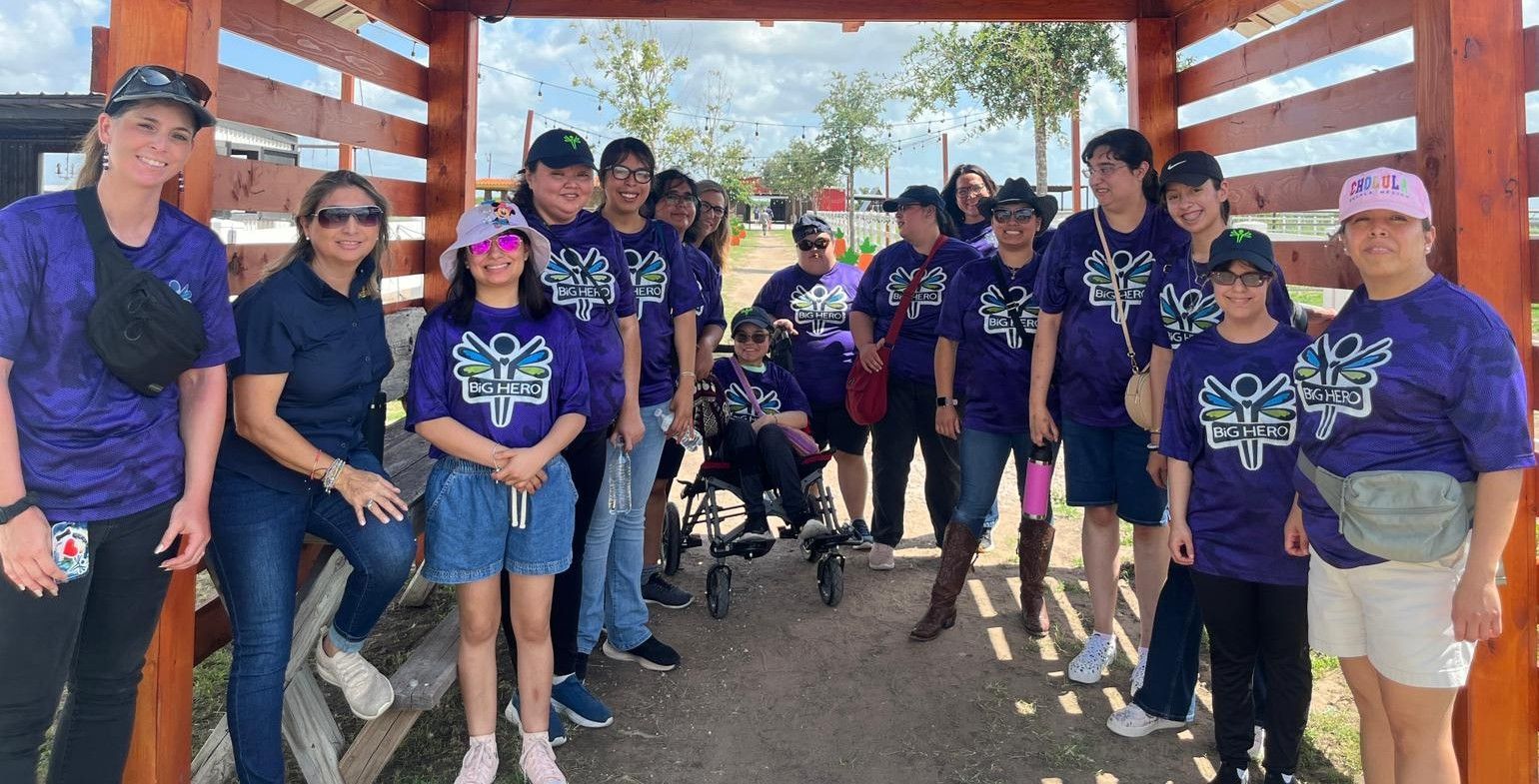Group of people in matching purple shirts, posing under a wooden archway, smiling in outdoor setting.