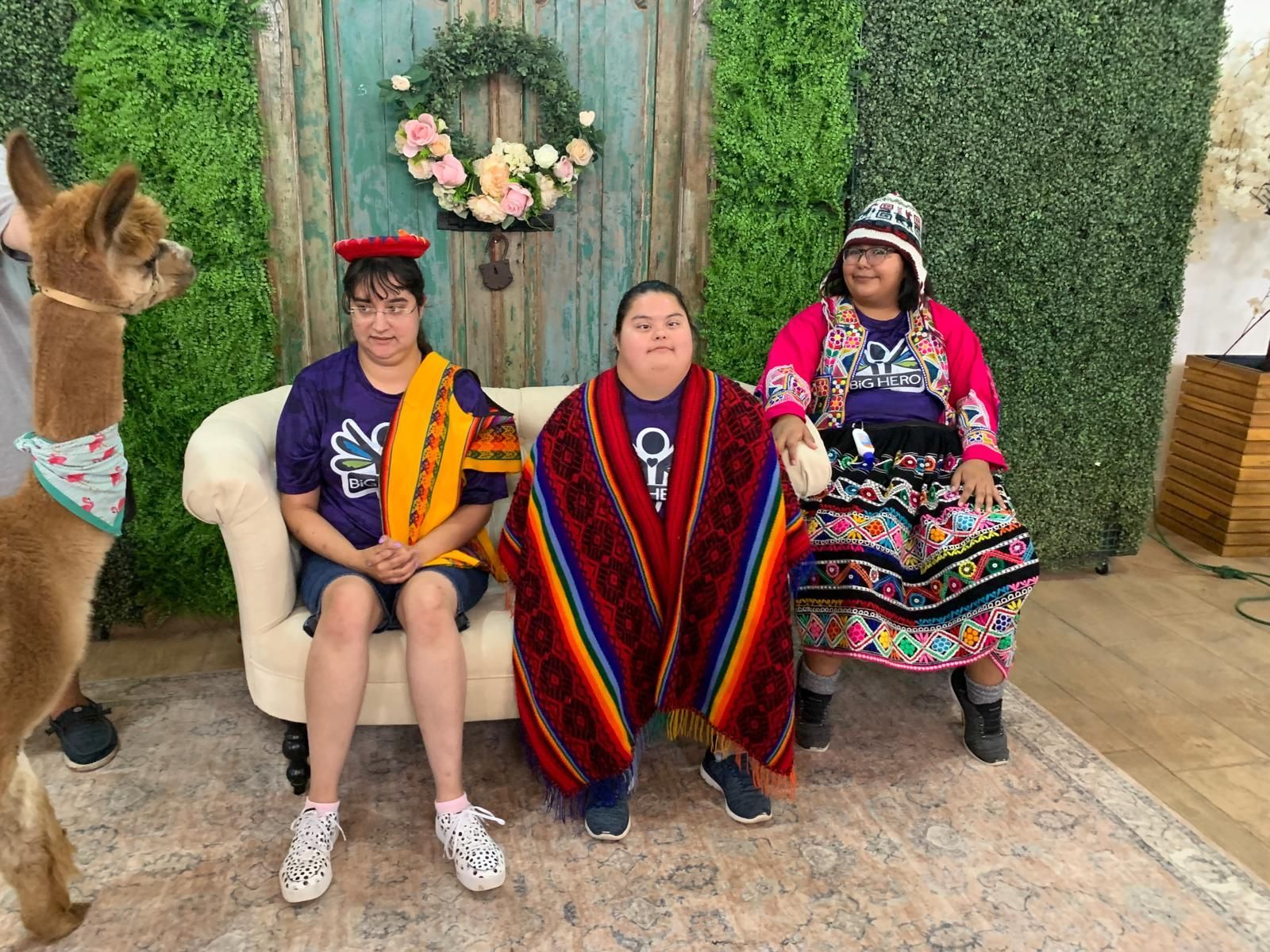 Three people in cultural attire with an alpaca posing on a couch in front of a green wall.