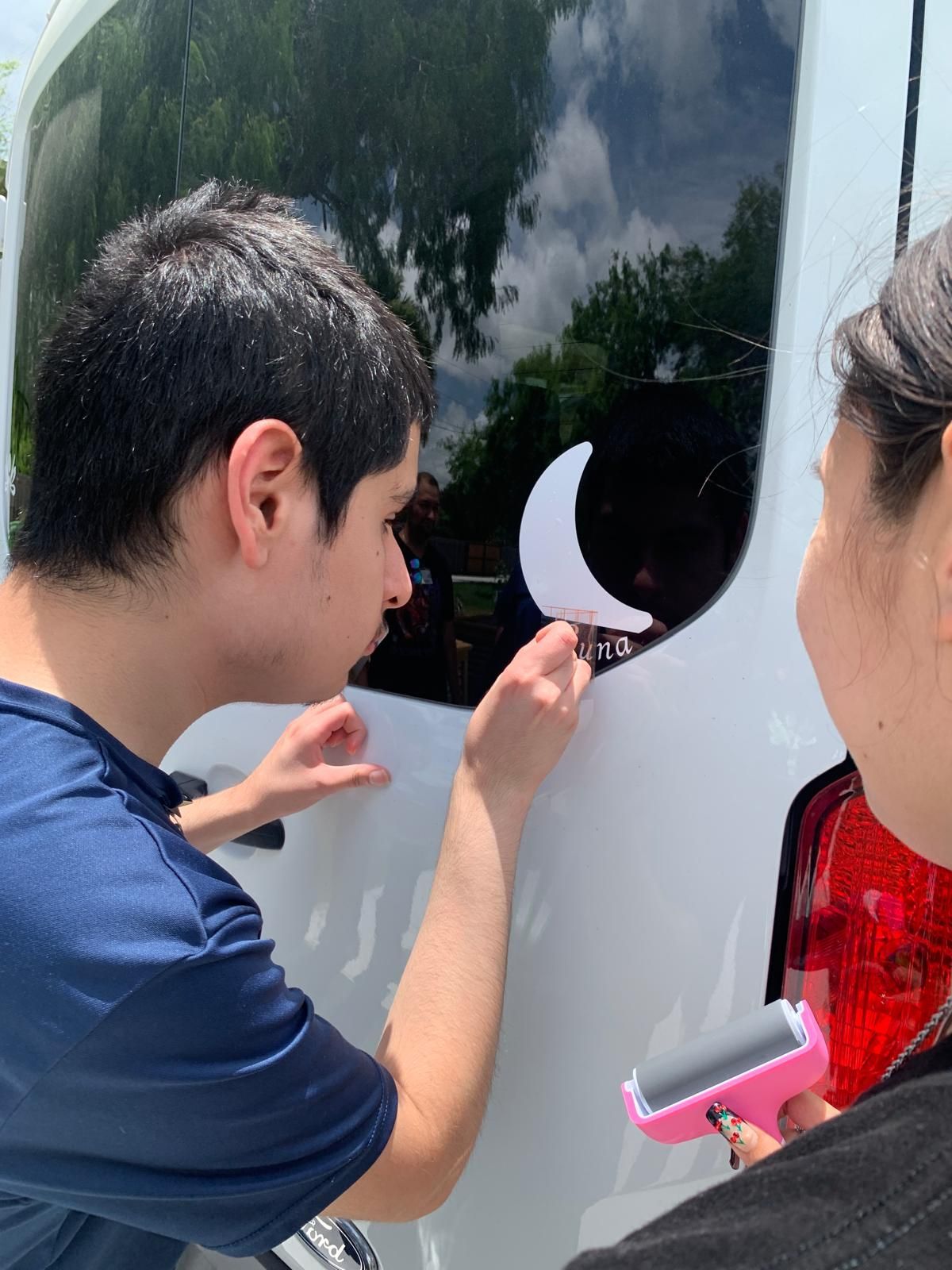 Two people applying a white crescent moon sticker to a van window; one holds a pink roller.