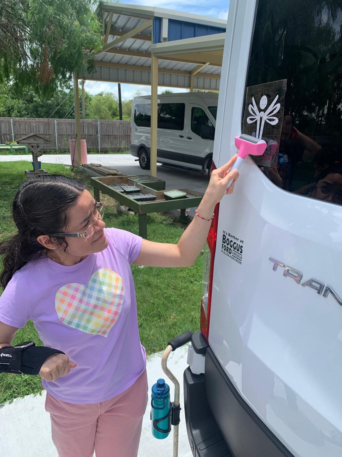 Woman with arm brace places a pink object on a van's window. She wears a purple shirt with a heart and pink pants.