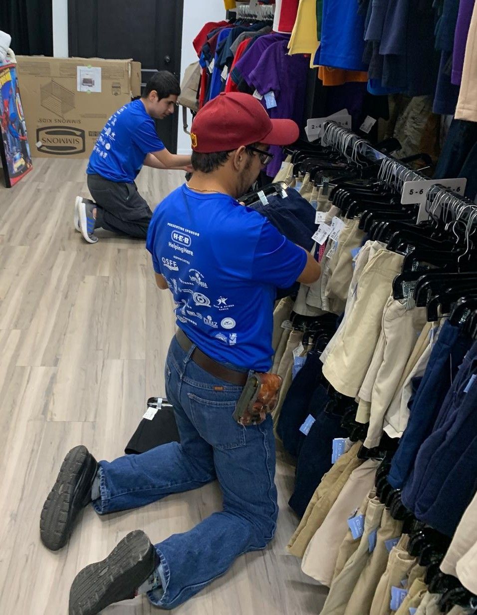 Two men kneeling, organizing clothing racks in a store. One wears a red cap, blue shirt, and jeans.