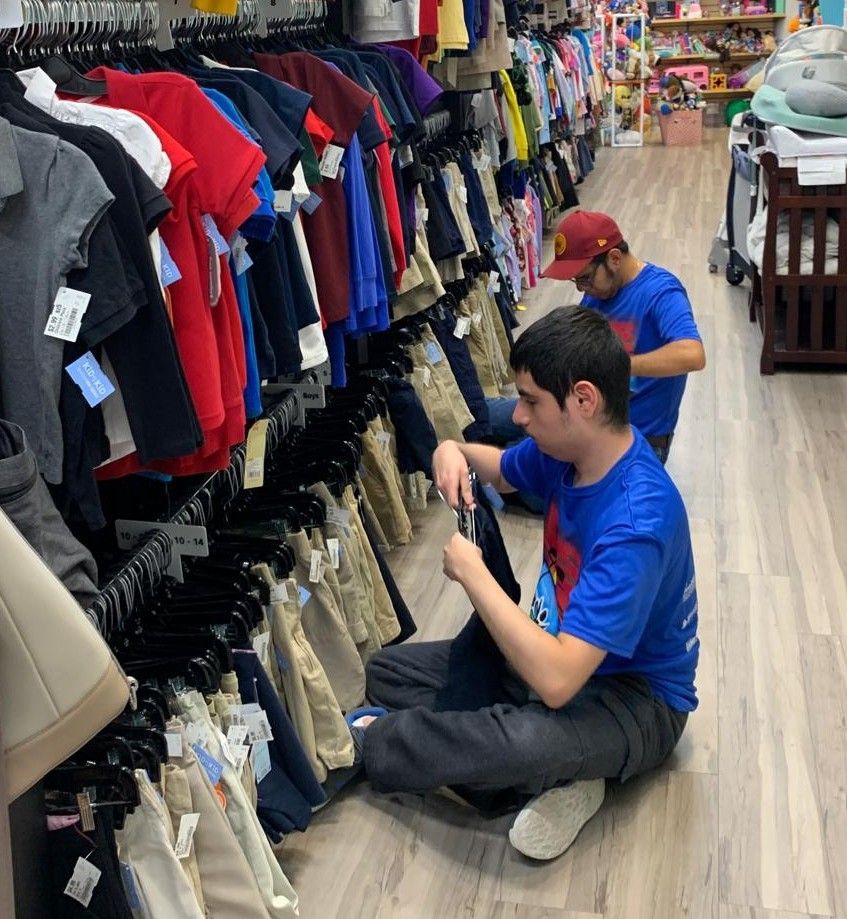 Two men in a clothing store, sorting through pants. One is on the floor, another is behind him.