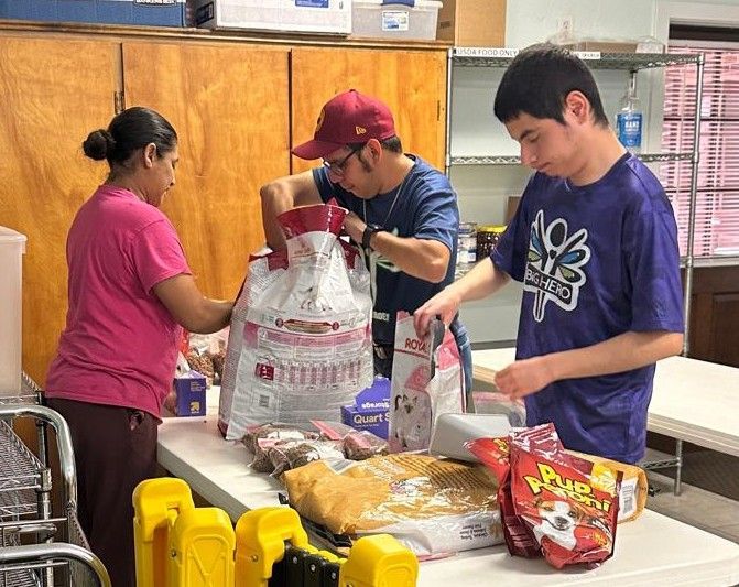 Three people packing pet food at a table. Woman, two men, various bags & boxes. Brown room, white table.
