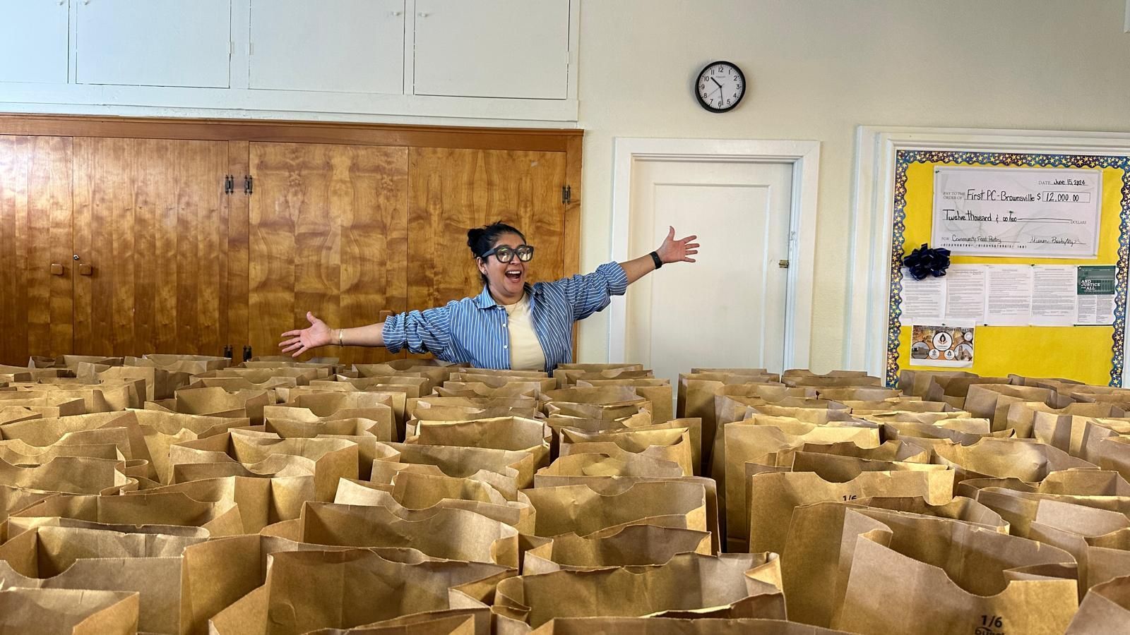 Woman in a room surrounded by brown paper bags, arms outstretched in a happy gesture.