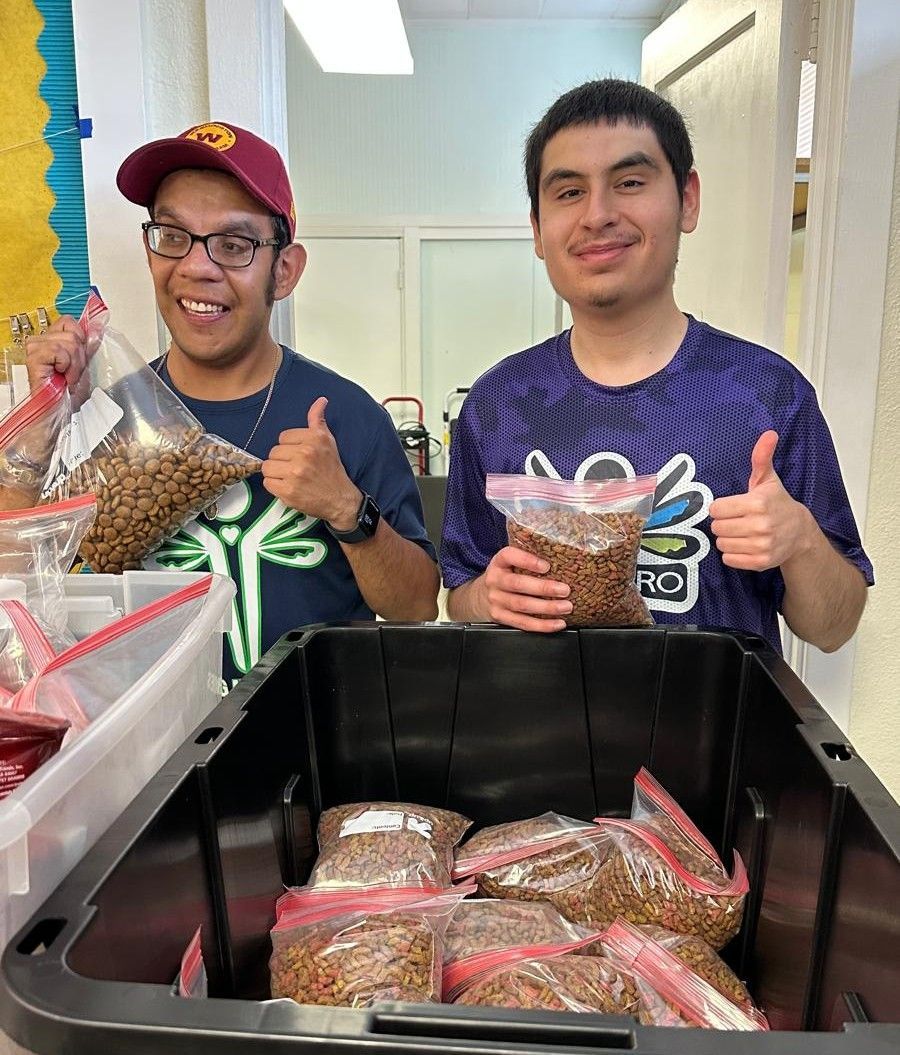 Two men smiling and giving thumbs up, holding bags of food. They are standing next to a bin of filled bags.