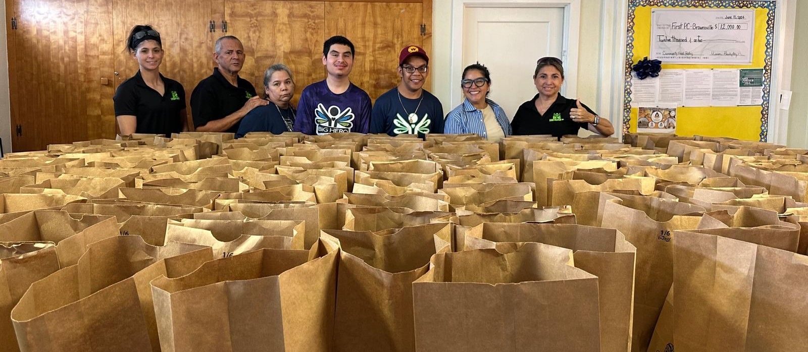 People standing behind many brown paper bags, possibly distributing food.