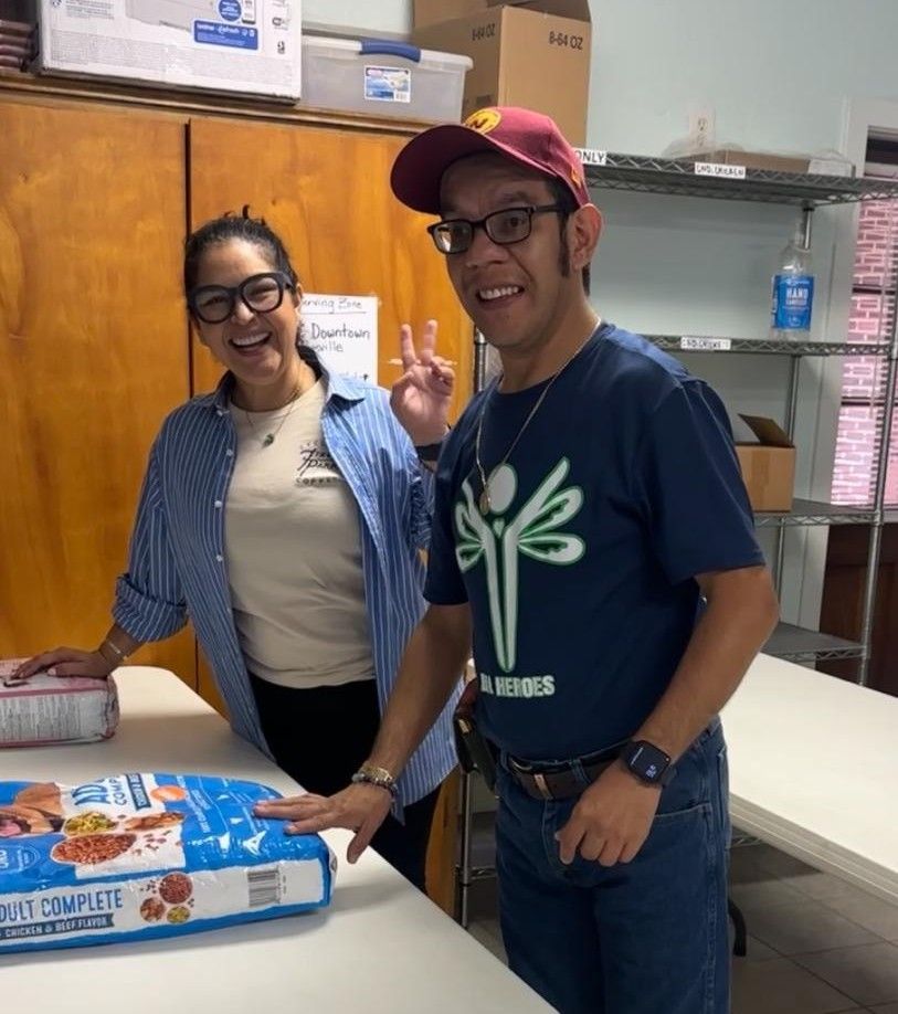 Two people smiling next to a bag of dog food in a pantry. One person gives a peace sign.