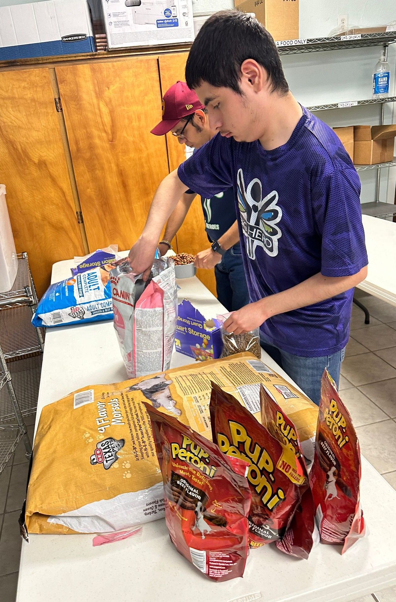 Two people packing pet food bags at a table indoors; one wears purple and one a red cap.