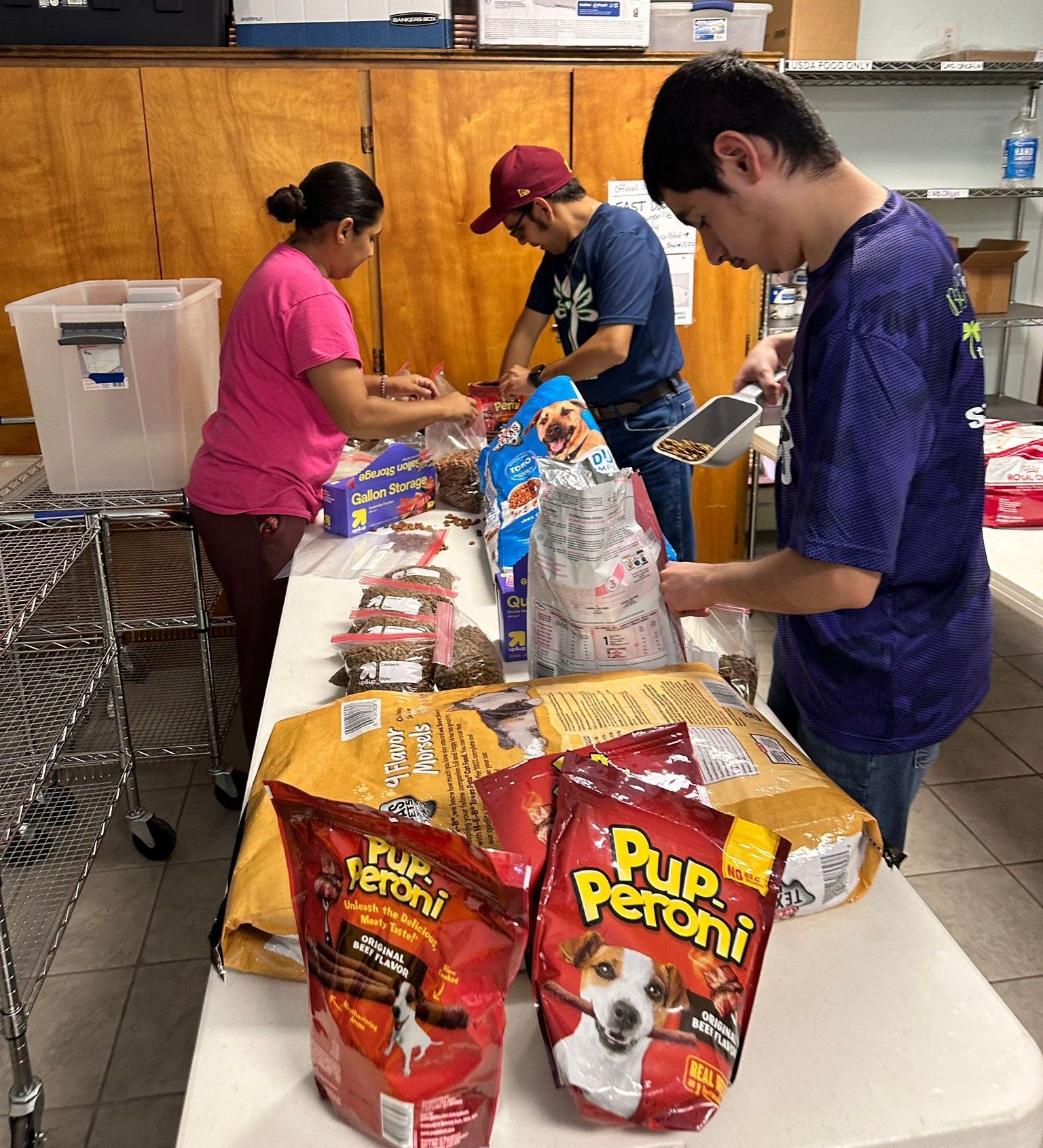 People packing pet food into bags at a table. Boxes and bags of food are visible.