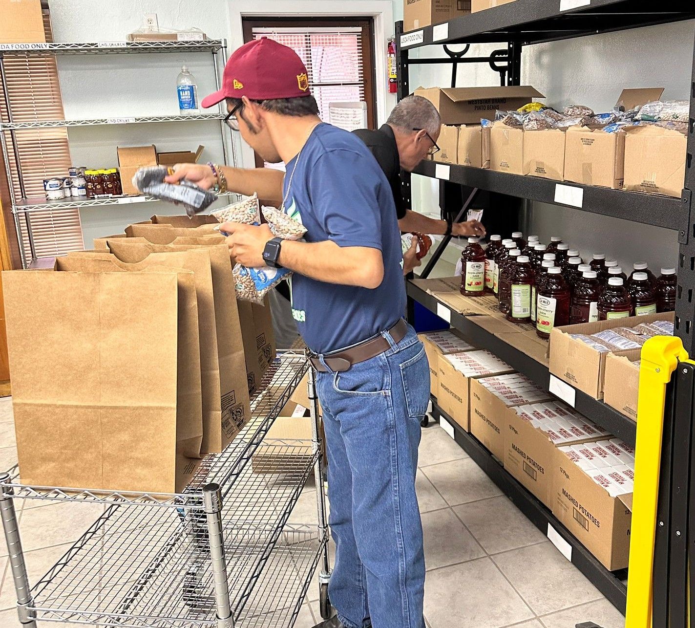 Two people in a pantry, scanning items into paper bags. Shelves hold food in boxes and jars.