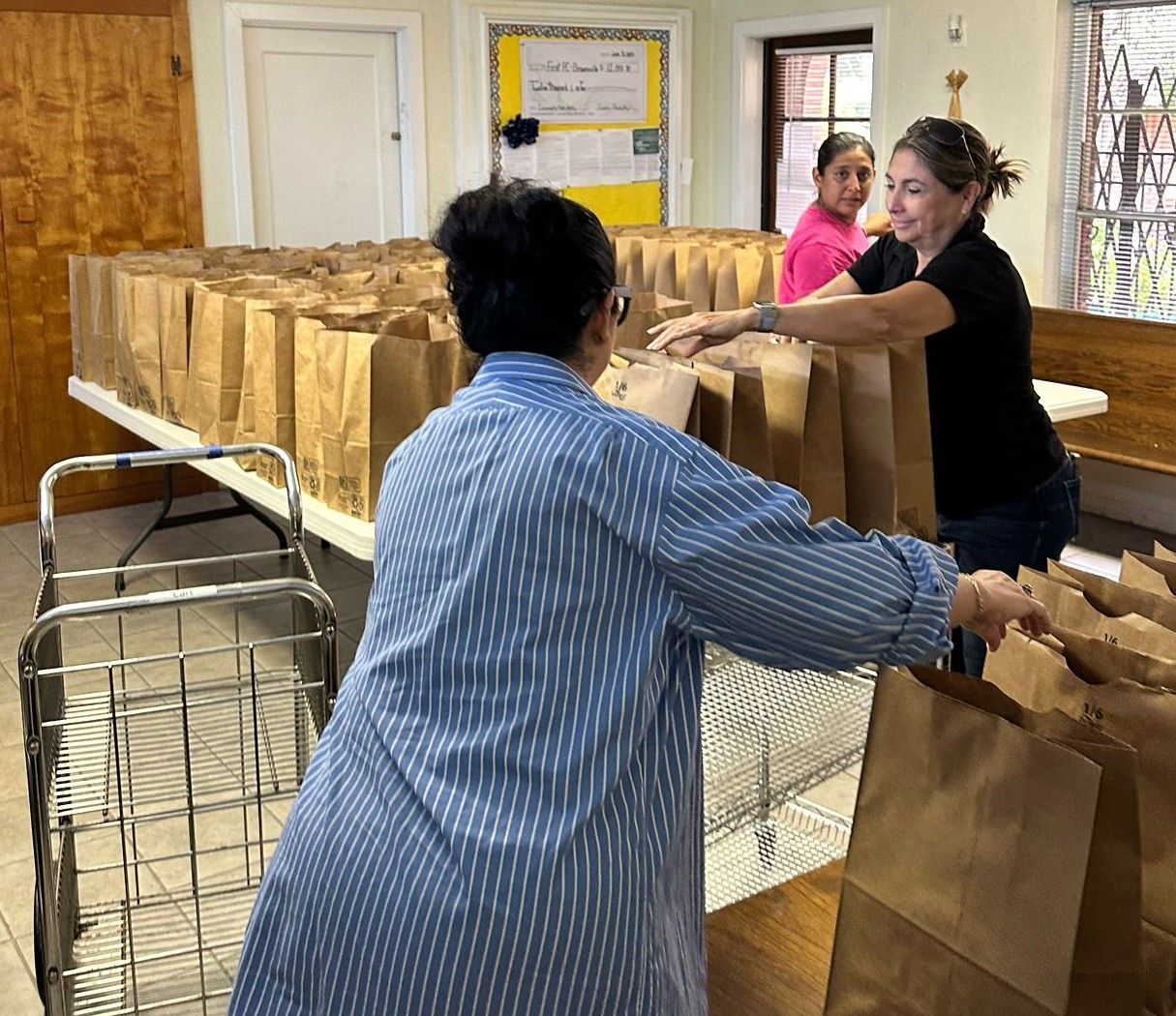 People packing brown paper bags at a table, likely food for distribution. A woman pushes a cart nearby.