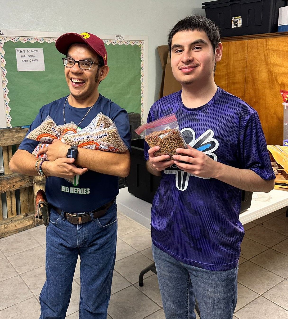 Two men holding bags of nuts, smiling. One wears a cap and blue shirt, the other a purple shirt.