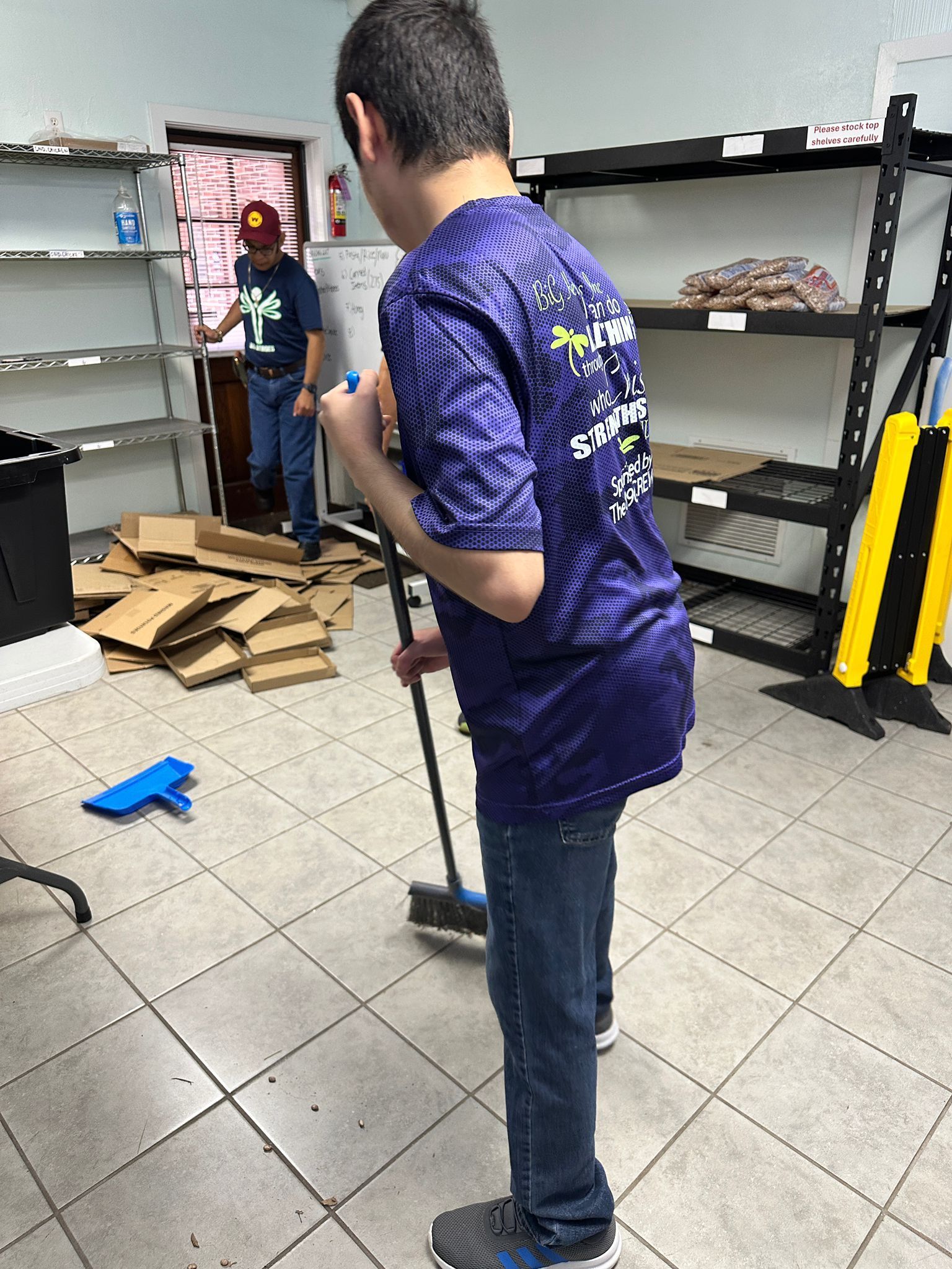 Young man sweeping a tiled floor, another person in background, warehouse setting.