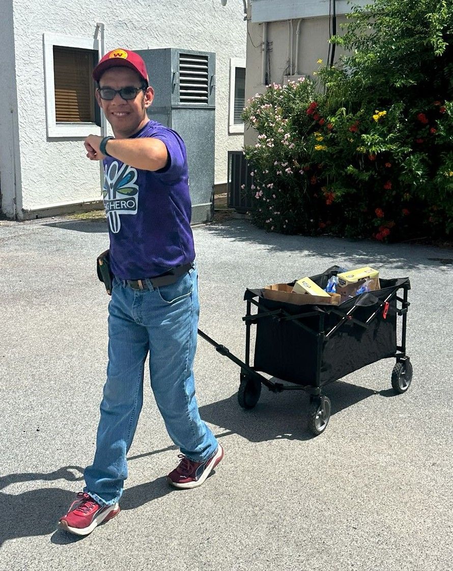 Man pulling a wagon, pointing forward. He wears a purple shirt, jeans, and a hat. Outdoors in sunny setting.
