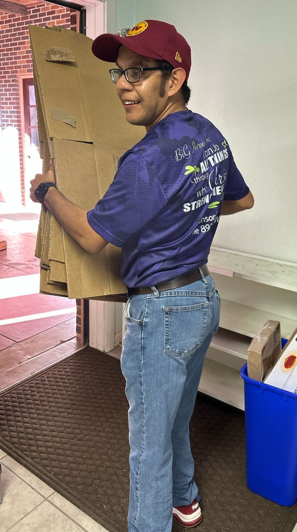 Person in maroon cap and blue shirt carrying cardboard, smiling.