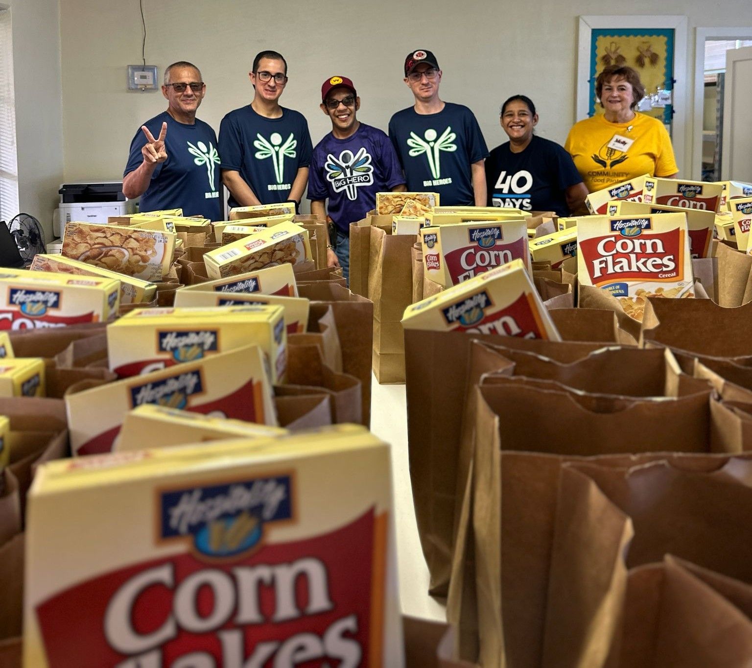 Volunteers packing food boxes. Corn flakes boxes and brown paper bags on a table. People smiling.