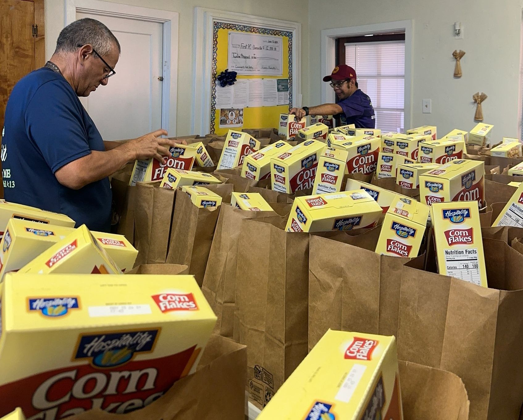 Two people packing groceries, including cereal boxes, into brown paper bags at a food bank.