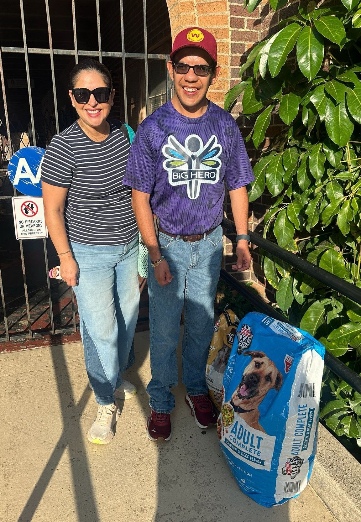 Two people stand outside, one holding a dog food bag.  Woman in striped shirt, man in tie-dye and cap, near a gate.