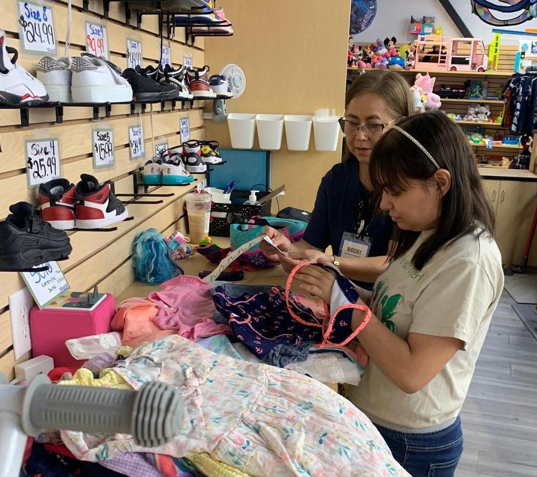 Woman and girl shop for clothes in a brightly lit store. Sneakers are displayed on a shelf.