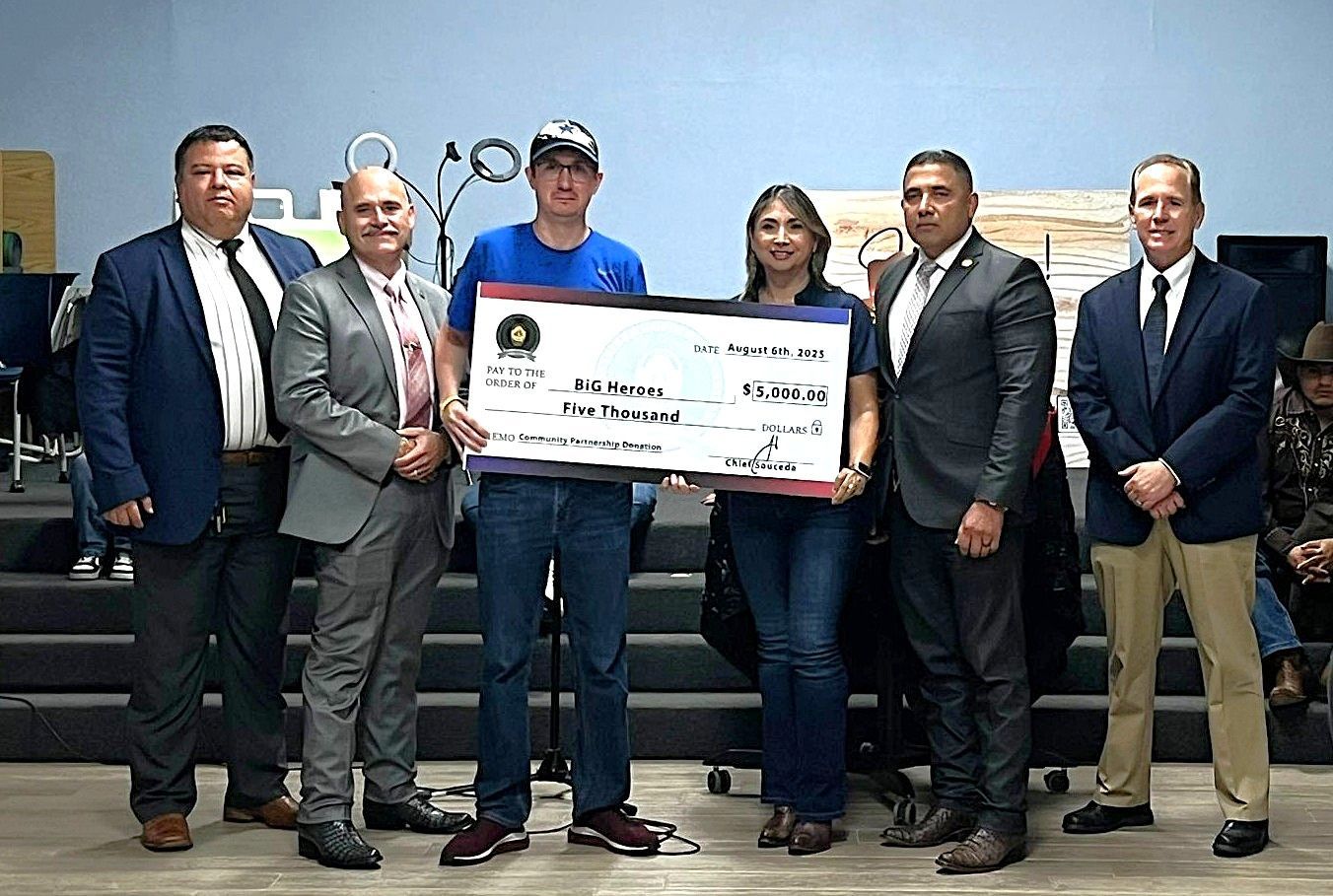 Group of six people holding large check on stage. Dark suits, jeans, casual attire.