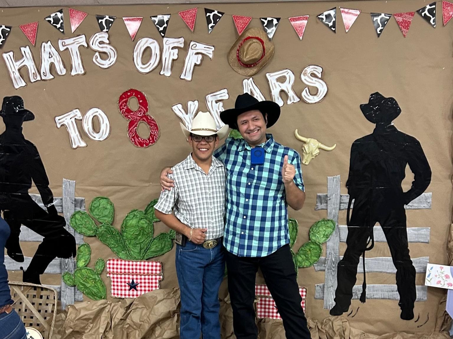 Two men in cowboy attire pose in front of a western-themed backdrop. One gives a thumbs up.