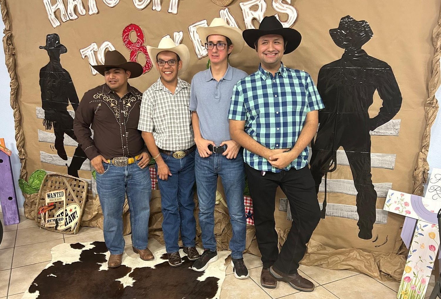 Four people in cowboy hats and outfits pose in front of a western-themed backdrop.