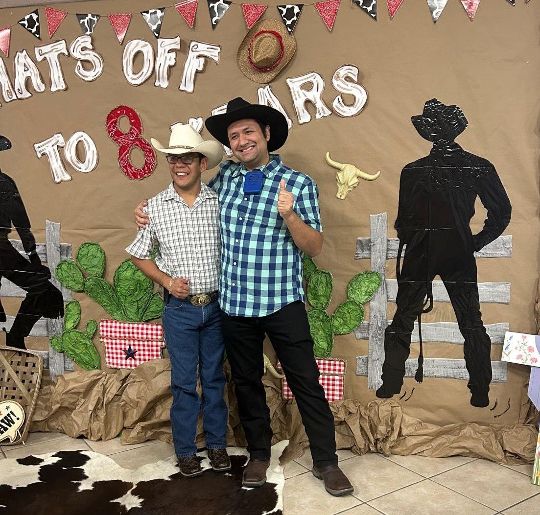 Two men in cowboy attire posing in front of a western-themed backdrop. One man gives a thumbs-up.