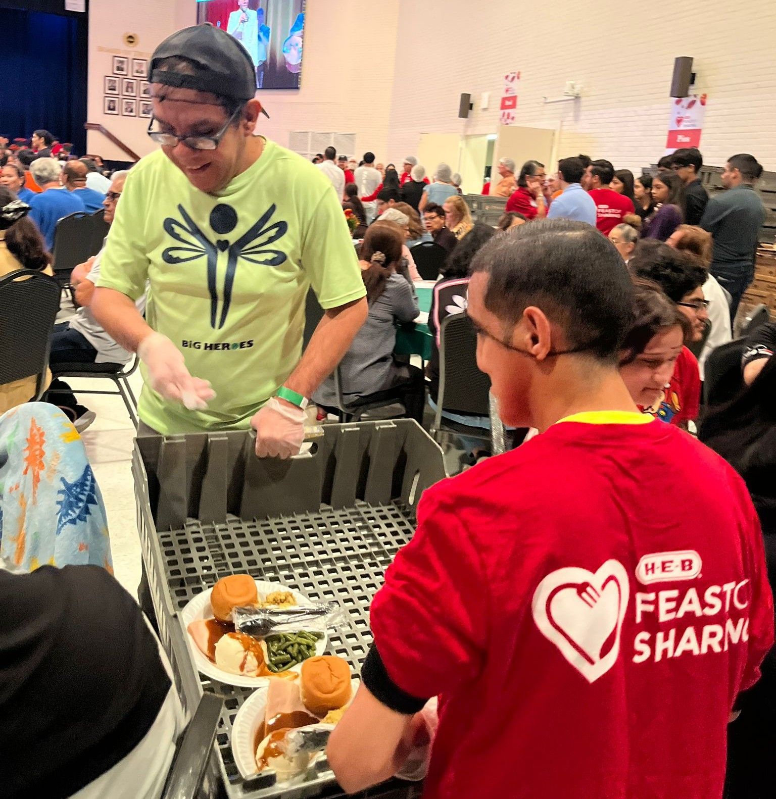 People serving food at an event. A person in a green shirt places food on plates. Others in red shirts also serve. Large gathering.