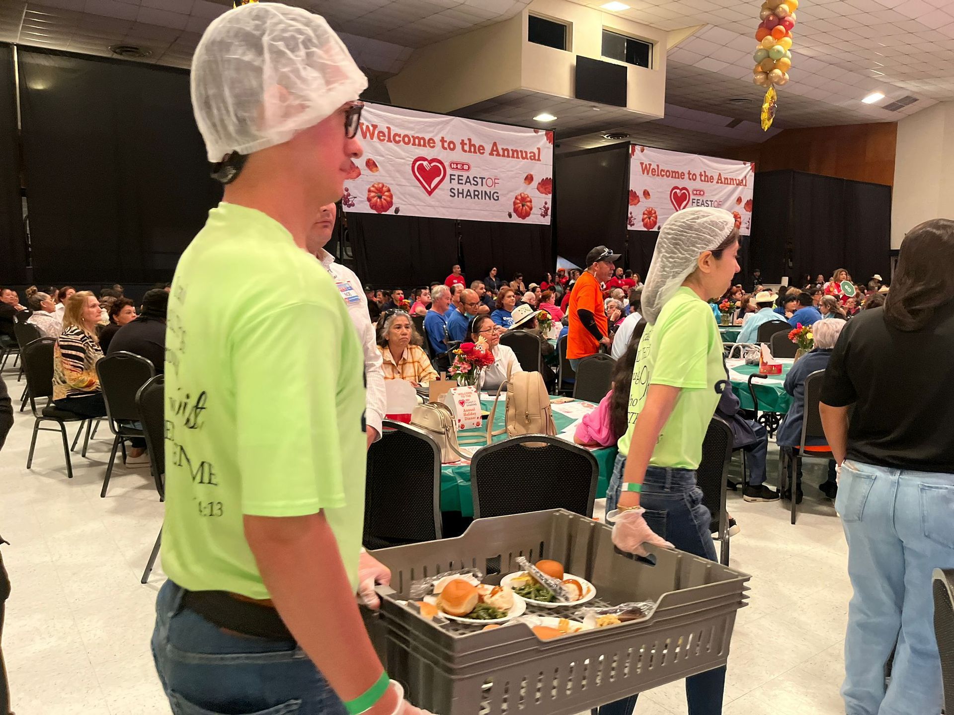 Volunteers serve food at a community event. People wear hairnets and carry trays in a crowded hall.