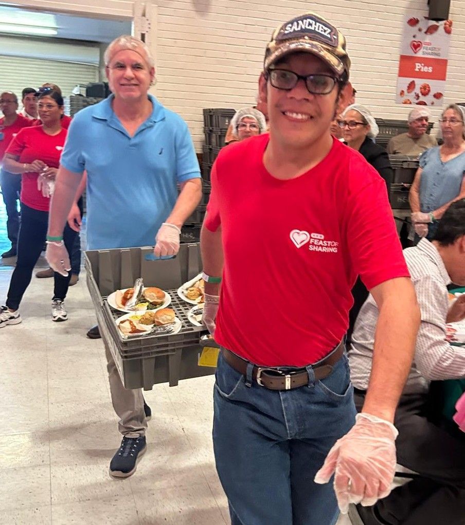 Volunteers serving food at a food bank. Man in red shirt carries tray of meals. Others in background.