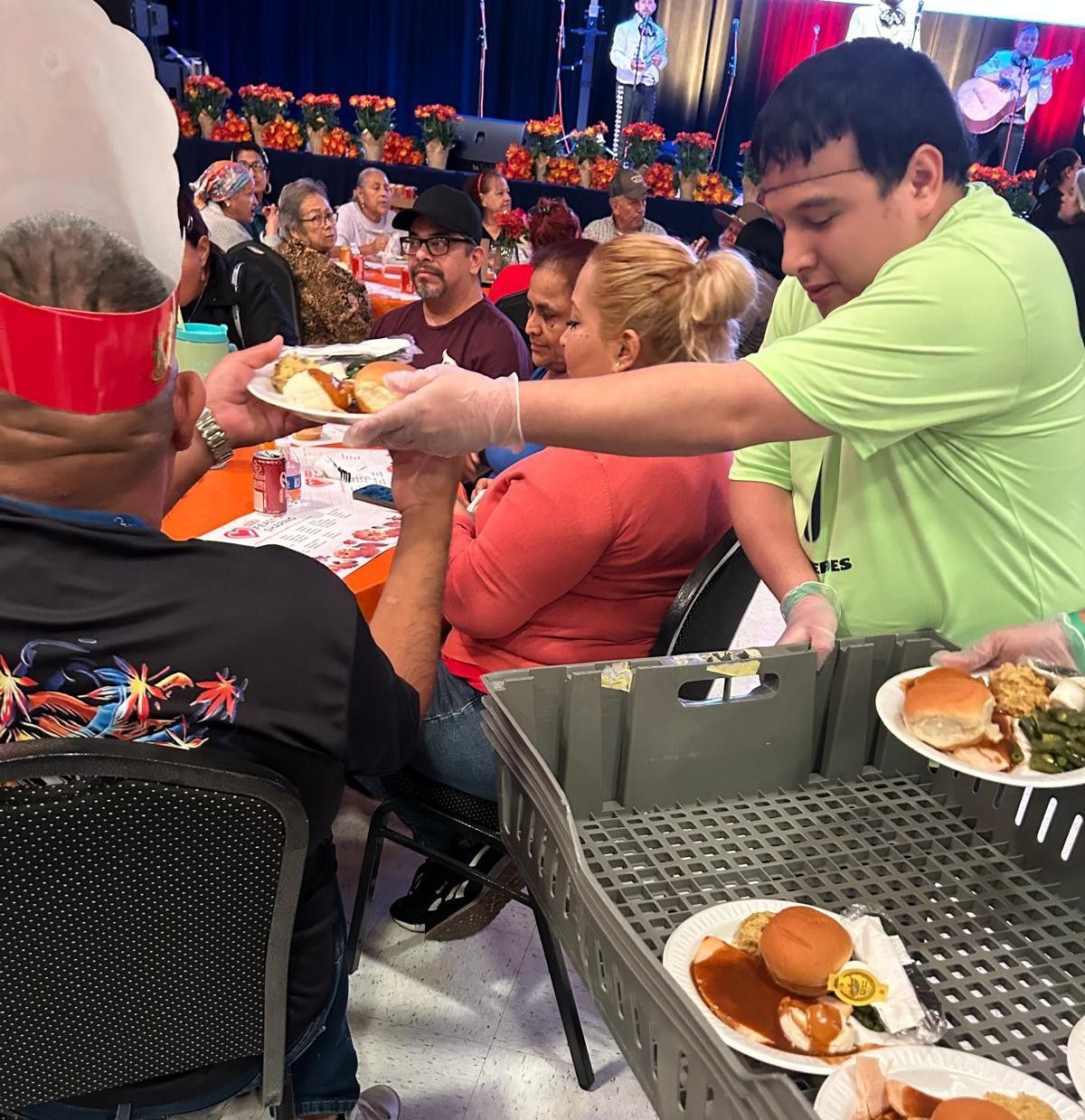 Man serves food to people at a community event. Plates of food are stacked in bins.