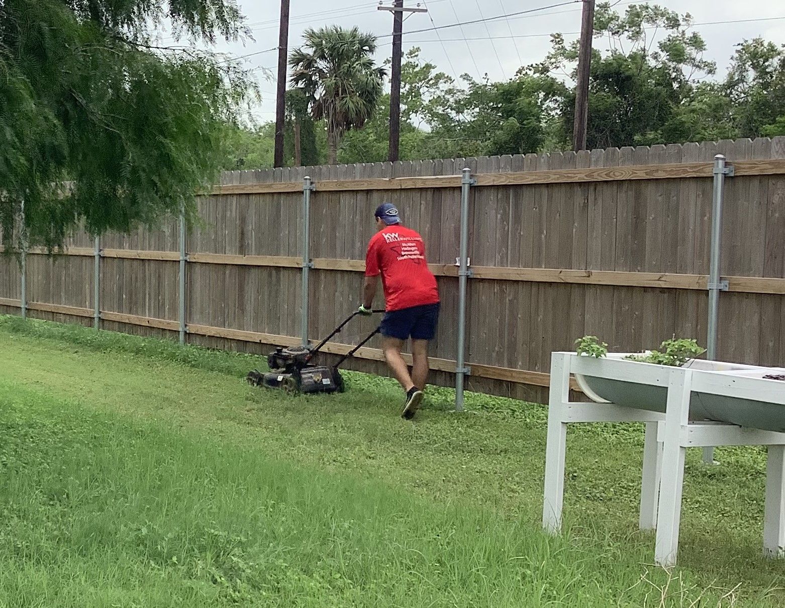 A man in a red shirt is mowing the grass in front of a wooden fence.