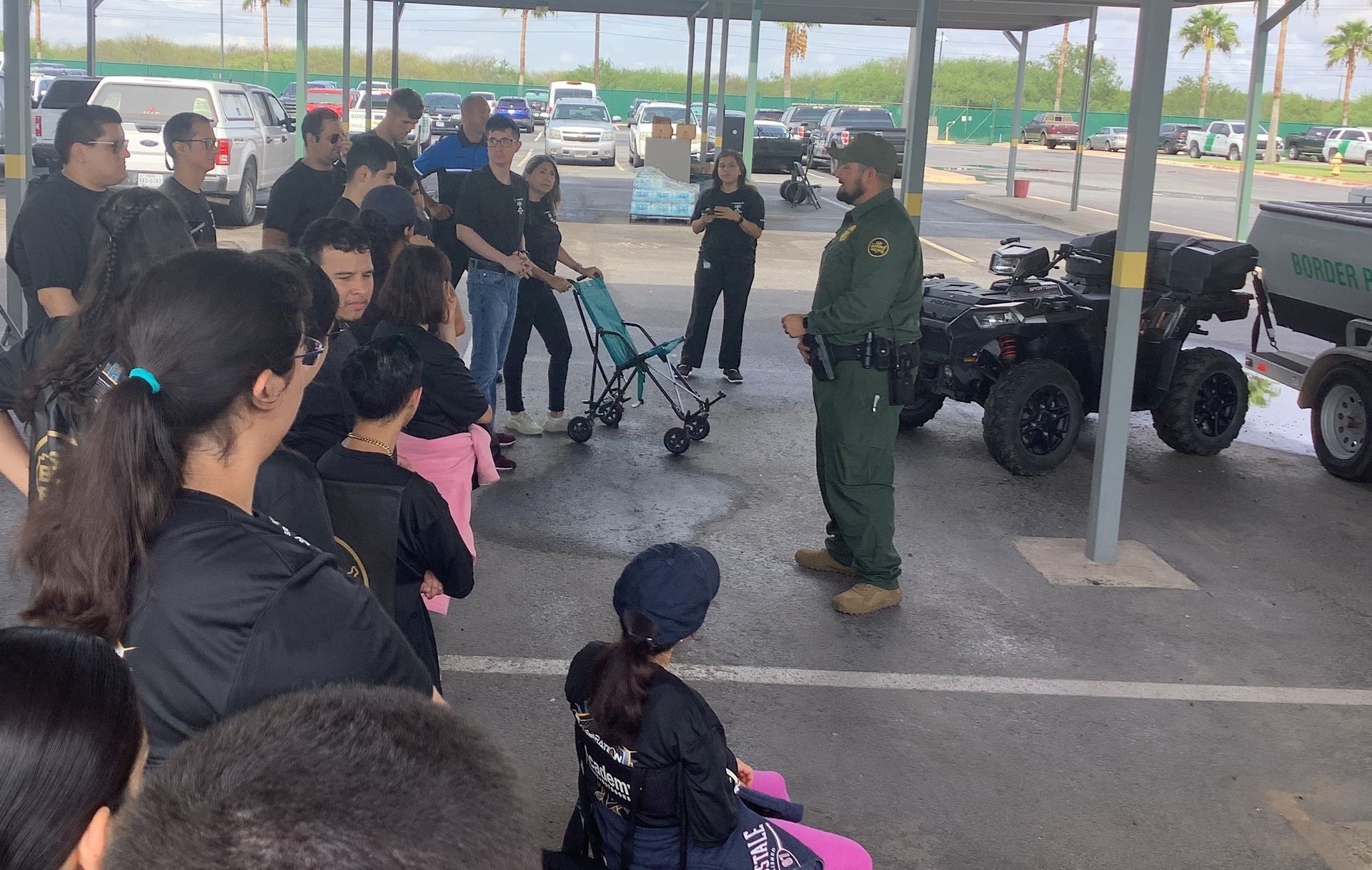 A man in a green uniform is talking to a group of people in a parking lot.