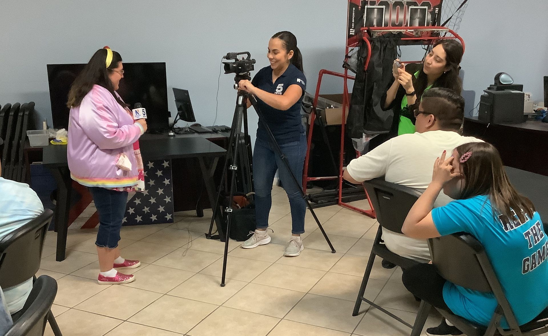 A group of people are sitting in chairs in a room with a woman standing in front of a camera.