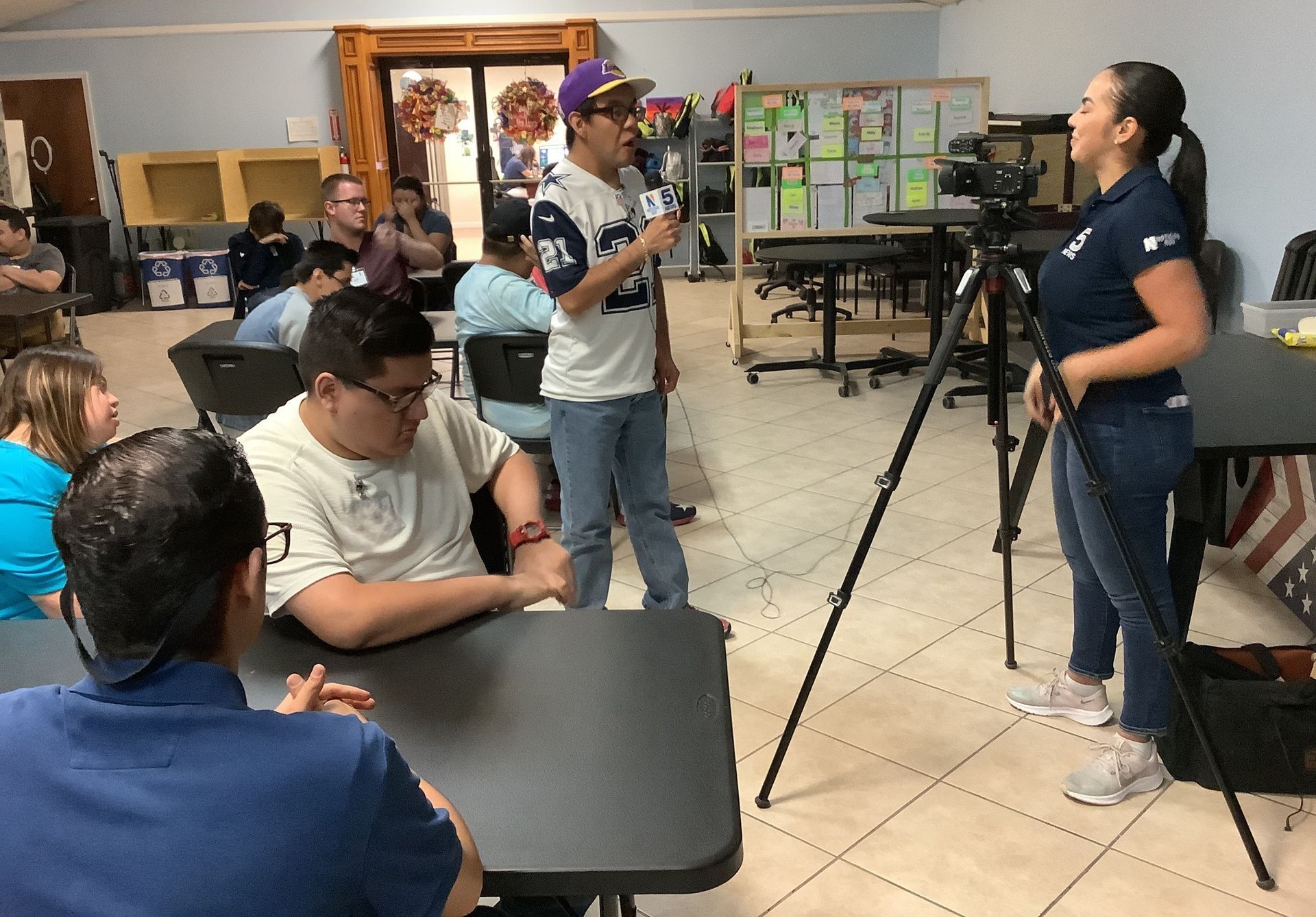 A group of people are sitting at tables and a woman is standing in front of a camera.