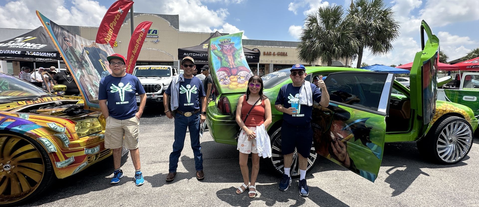 Four people pose with customized cars at an outdoor event, including one with open doors.