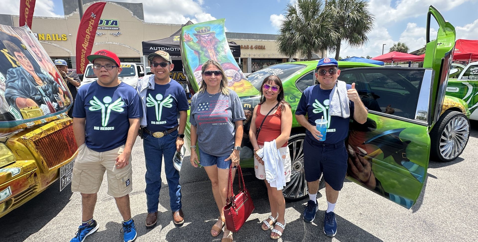 Five people pose by customized cars at an outdoor event. Brightly colored, sunny day.