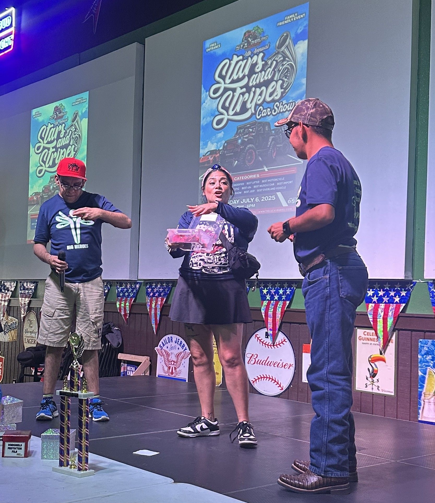 Three people on stage. Woman holding a container; men beside her, one gesturing. Event backdrop with patriotic theme.